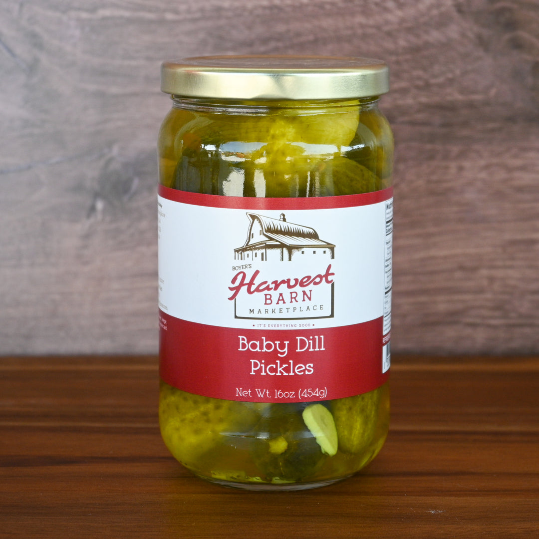 Jar of baby dill pickles with a Harvest Barn label on a wooden surface.