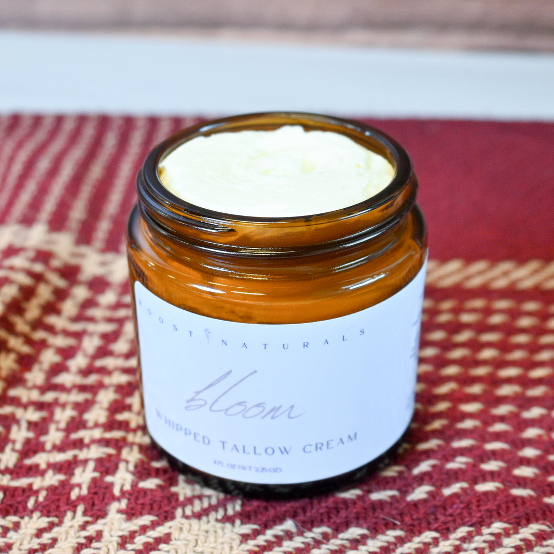 An open amber glass jar of East Naturals Bloom Whipped Tallow Cream, showing the pale yellow cream inside, set on a red and beige patterned woven mat.
